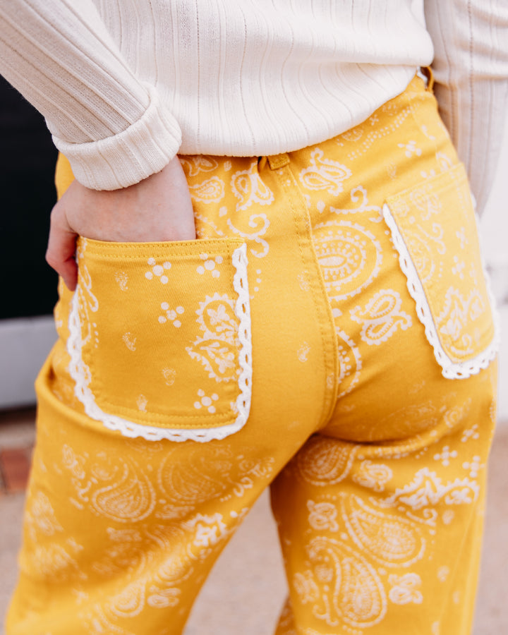 Close-up of a person wearing yellow bandana-print pants with white scalloped pocket trim, paired with a cream ribbed sweater.