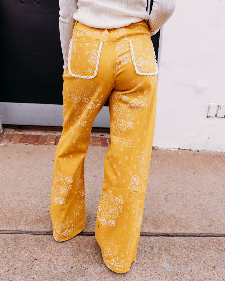 A person wearing mustard yellow wide-leg pants with a white paisley bandana print and white lace trim on the back pockets, standing outdoors on a sidewalk.