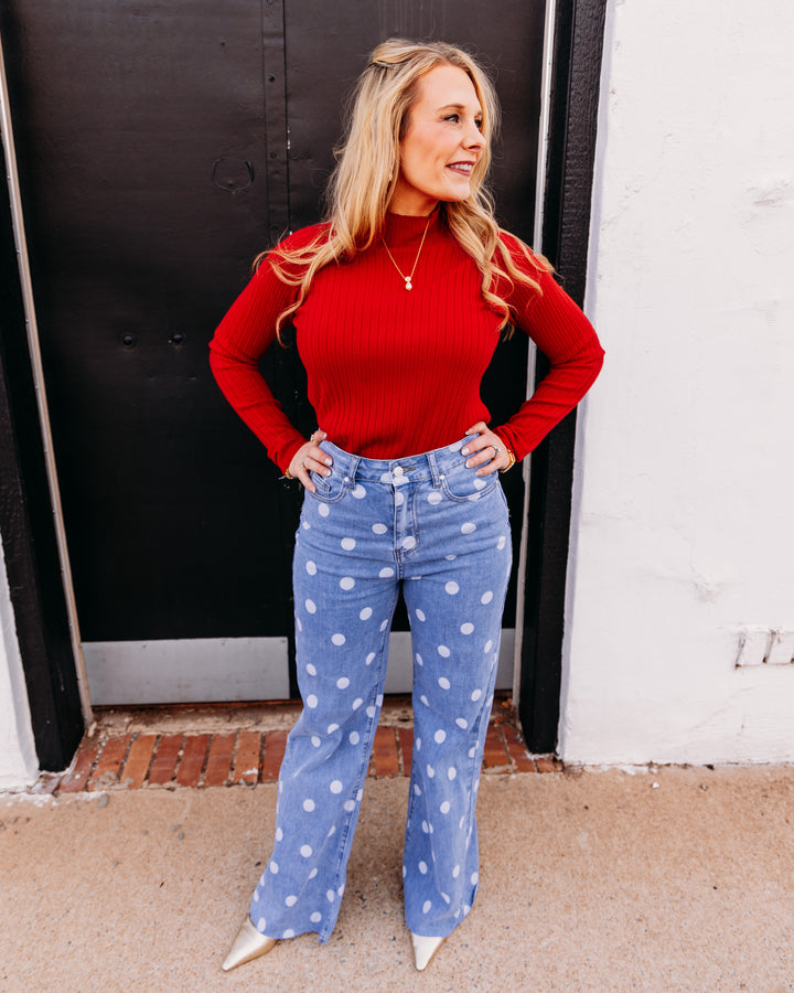 A woman stands outdoors in front of a black door and white wall, wearing a fitted red ribbed long-sleeve top and light blue wide-leg jeans with white polka dots. She is also wearing pointed silver heels and a delicate gold necklace.