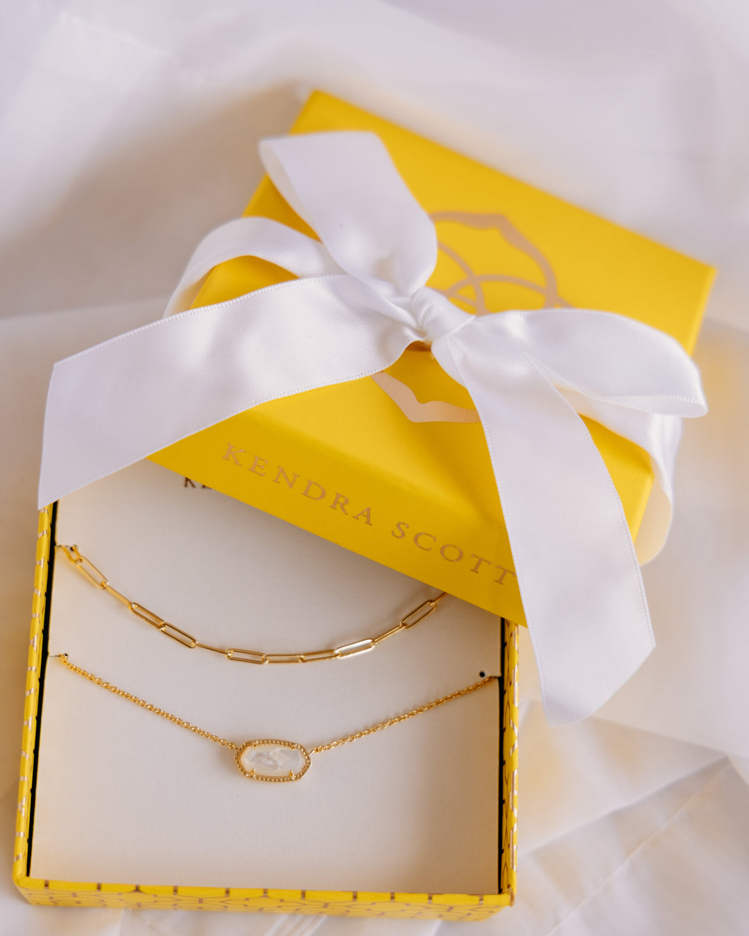 Two gold necklaces, one with an oval white stone and one with a link chain design, presented in a yellow jewelry box tied with a white ribbon. The brand name 'KENDRA SCOTT' is visible on the box.