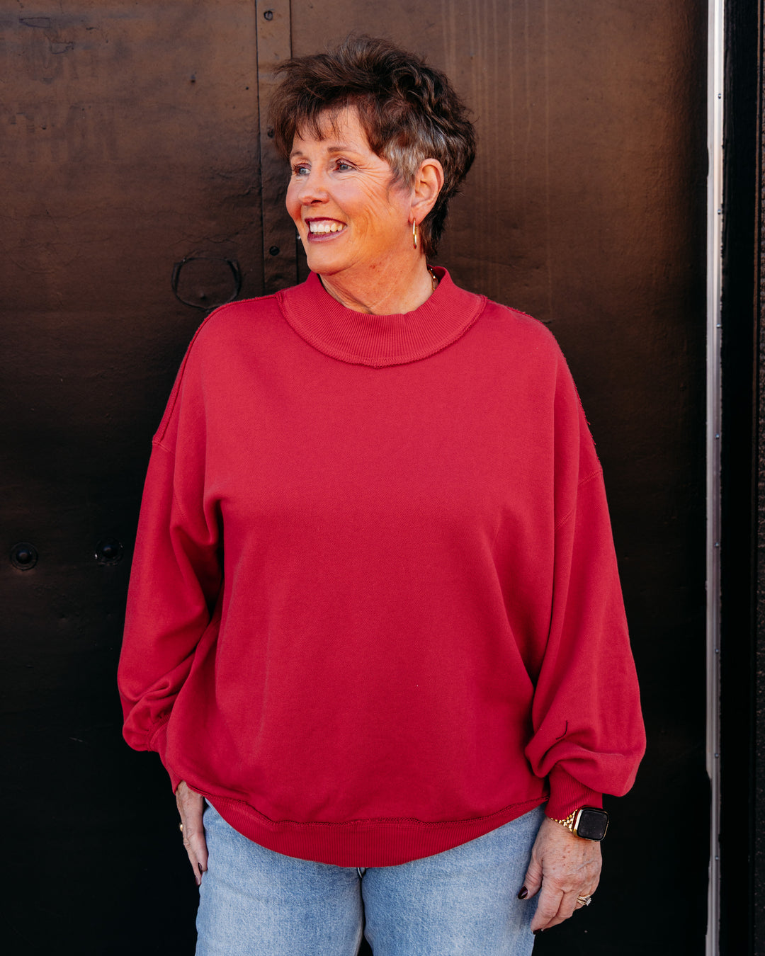 A smiling woman with short brown hair wearing a bright red mock-neck sweatshirt and light blue jeans, standing in front of a dark brown wooden door.