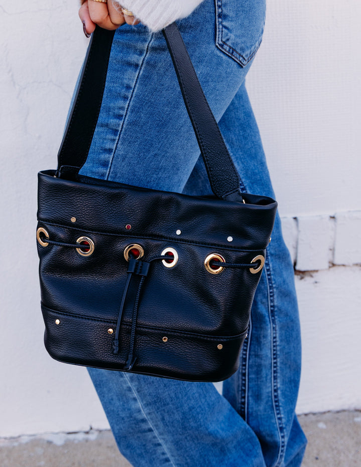 A person wearing blue jeans and a white sweater carries a black leather shoulder bag with gold-tone hardware against a white wall.