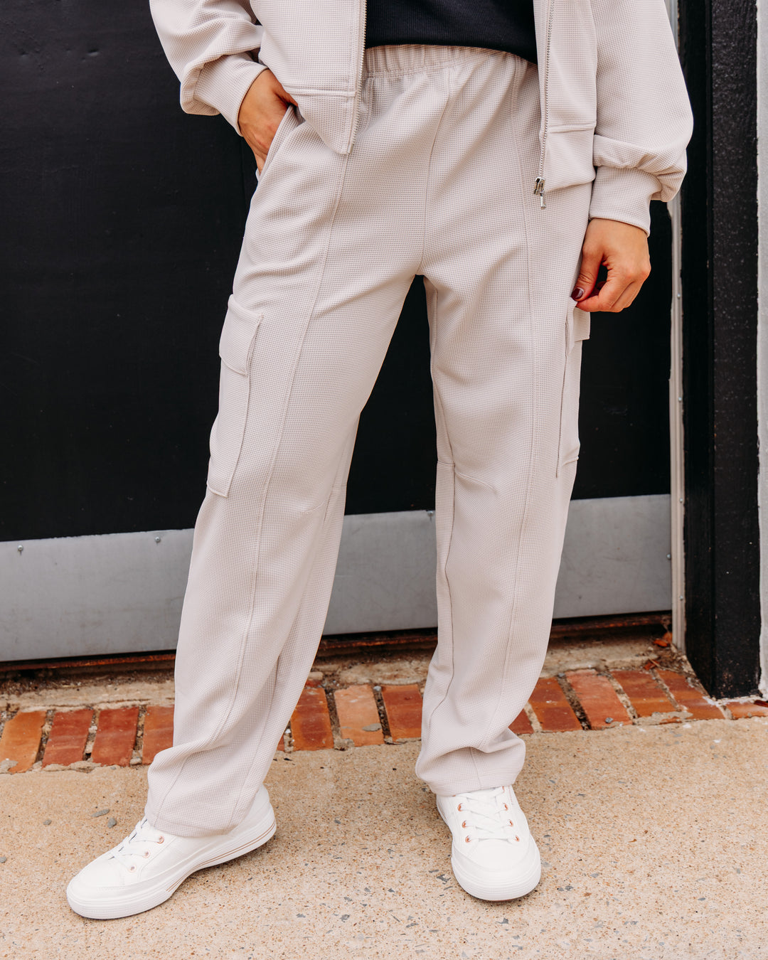 A person wearing light beige cargo jogger pants, a matching zip-up hoodie, and white sneakers, standing outdoors in front of a dark wall.