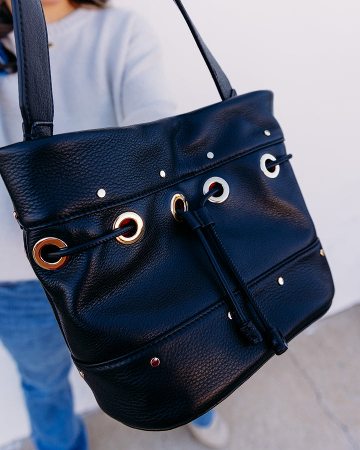A black pebbled leather shoulder bag with gold and silver-tone metal grommets and studs, shown being carried by a person wearing a light gray top and blue jeans against a plain light background.