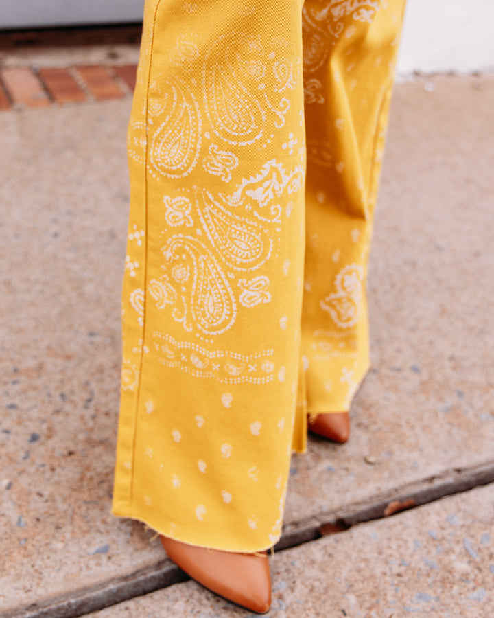 Yellow wide-leg pants with a white paisley bandana print, shown on a model wearing tan pointed-toe heels on a concrete sidewalk.