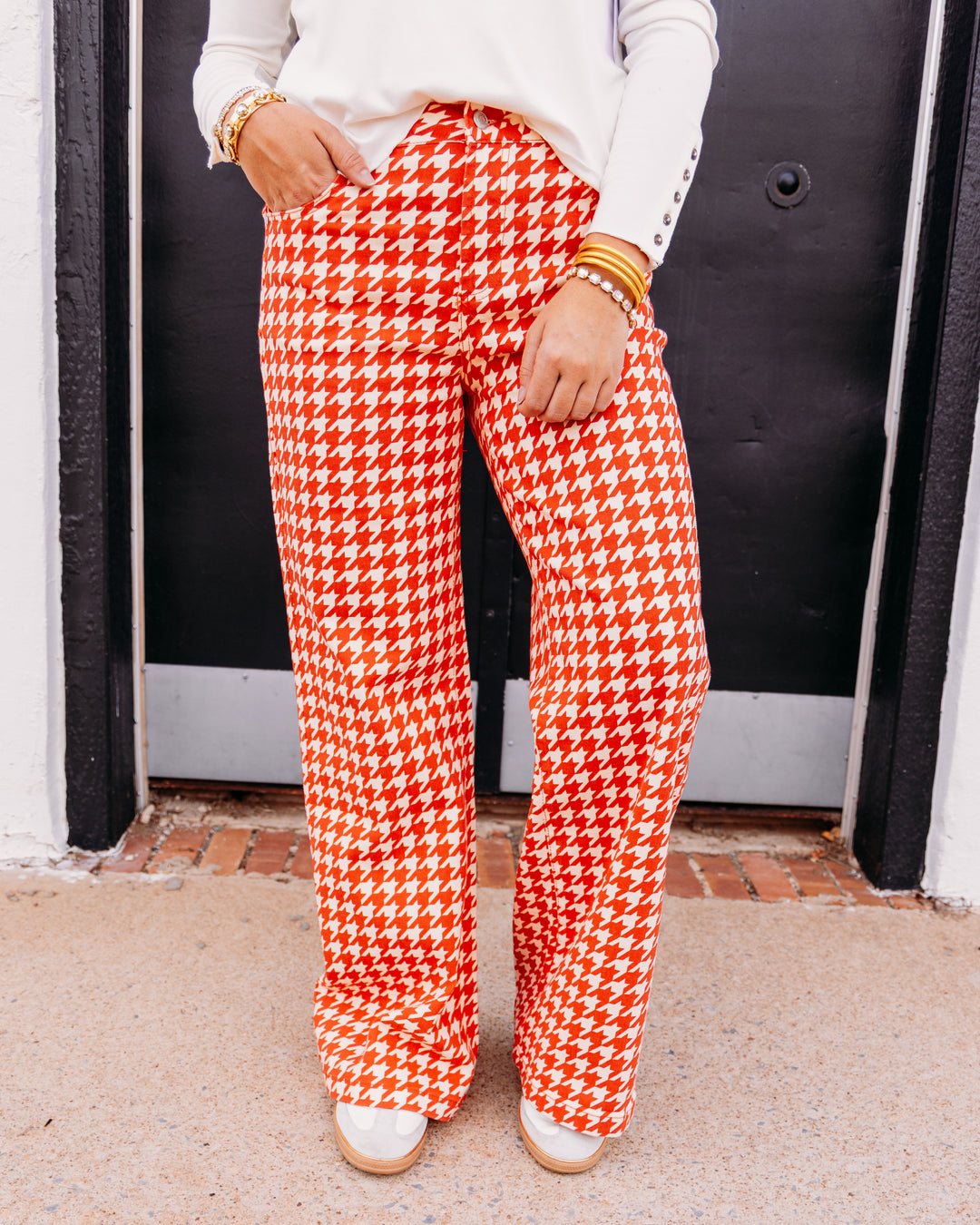A person wearing bright orange and white houndstooth wide-leg pants, styled with a white long-sleeve top and white shoes, standing in front of a black door.