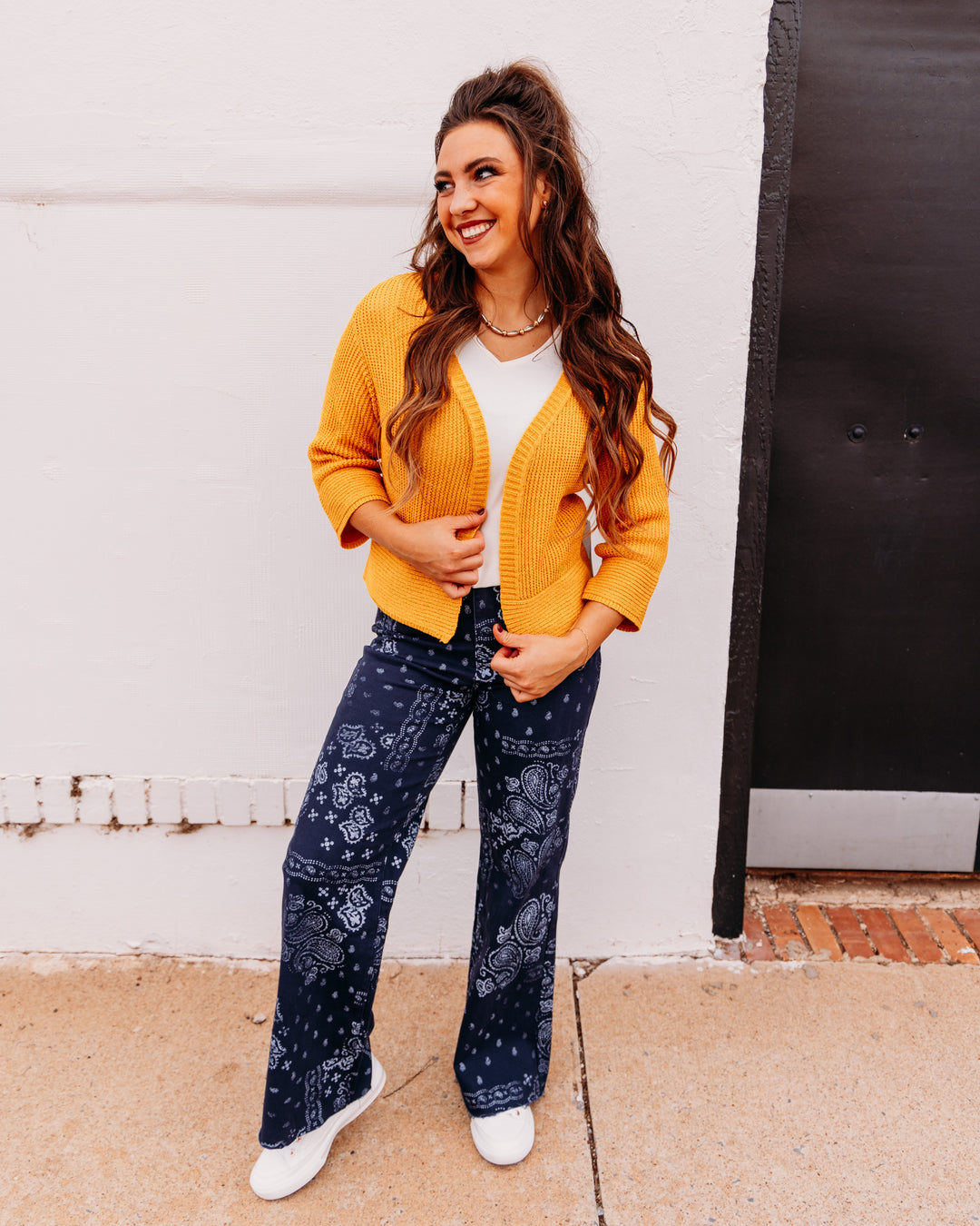 A woman modeling a mustard yellow cardigan over a white top and navy blue bandana print pants, standing outdoors against a white wall.