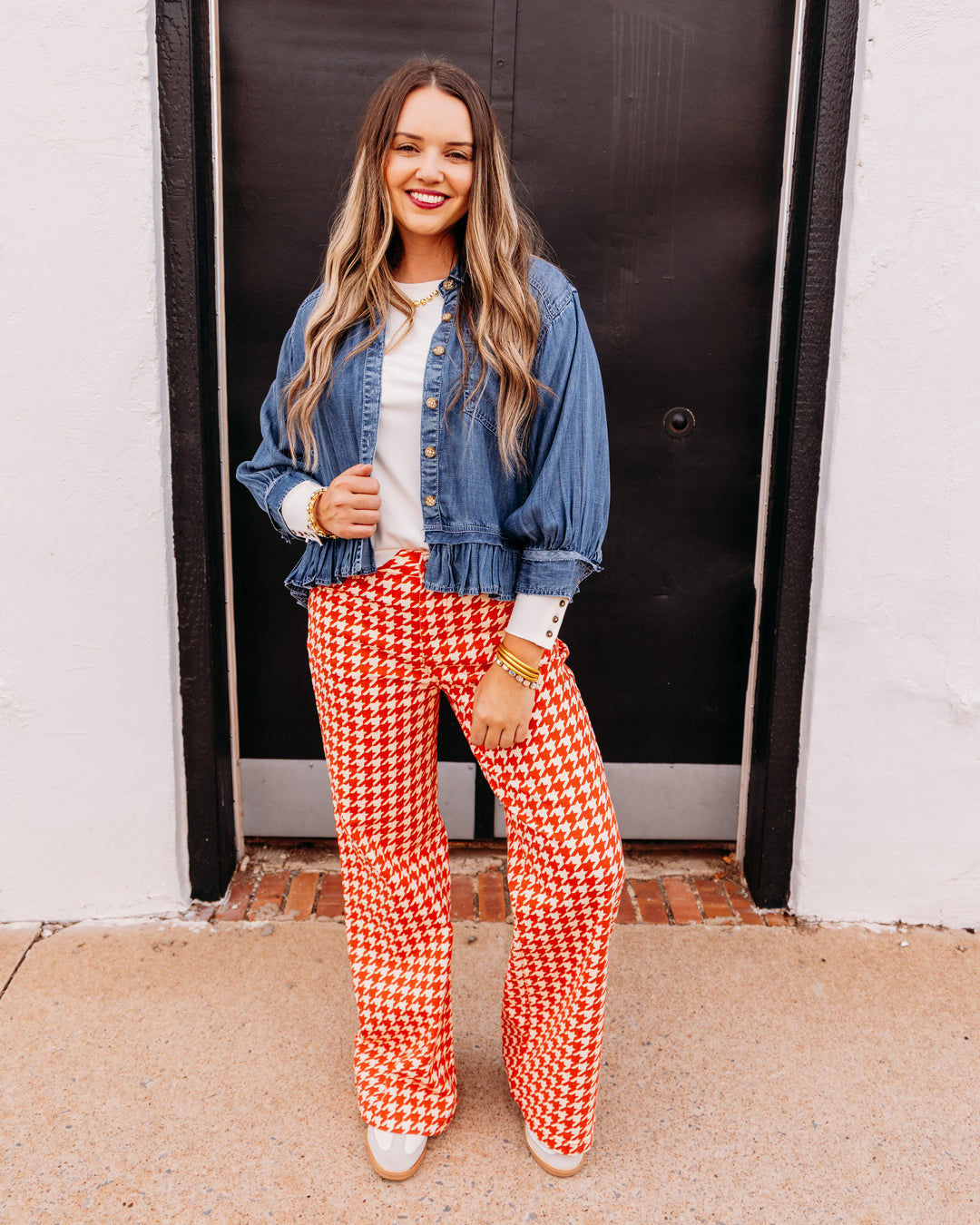 A woman stands in front of a black door wearing a blue denim jacket, a white top, and red and white houndstooth wide-leg pants.