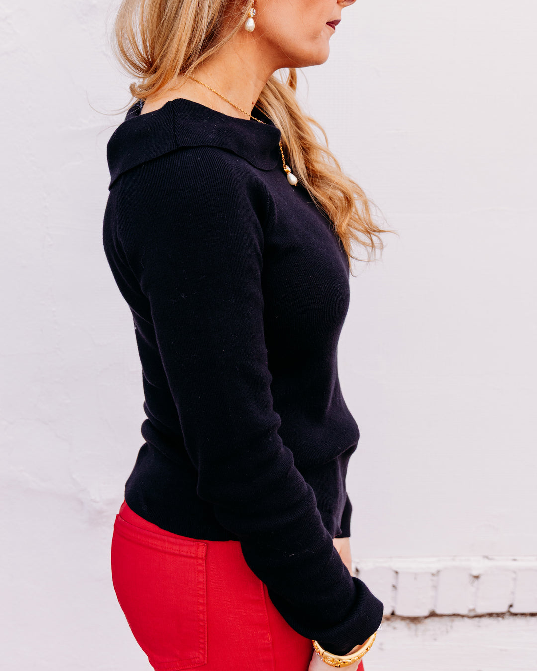 A woman shown in side profile wearing a fitted black long-sleeve top and bright red pants against a plain white wall.