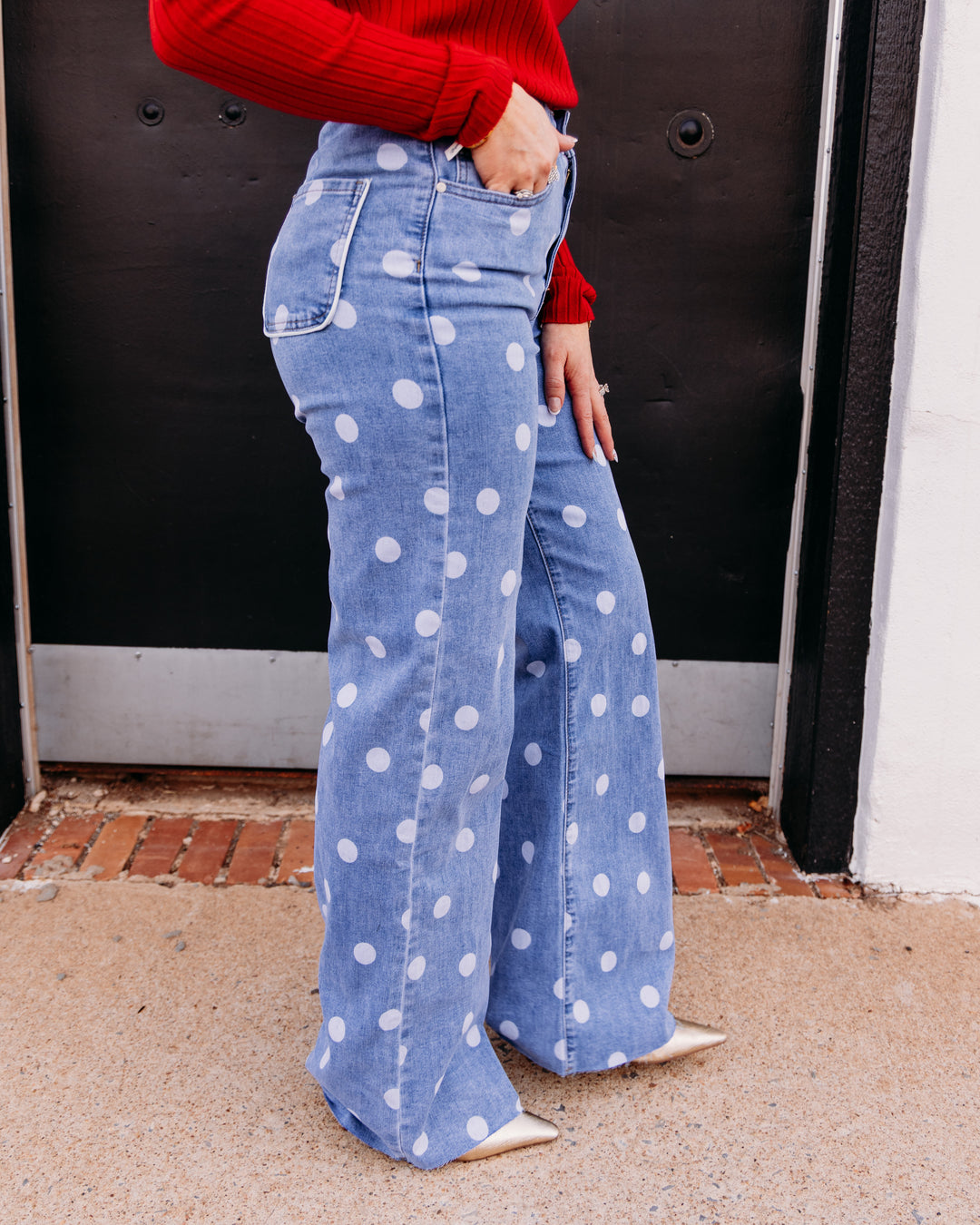 A person wearing light blue wide-leg jeans with white polka dots, paired with a red ribbed sweater and beige pointed-toe flats, standing in front of a black door.