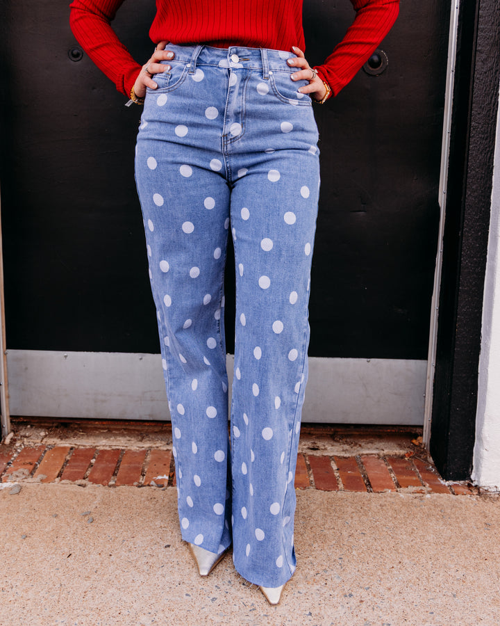 A person wearing light blue high-waisted flared jeans with large white polka dots, paired with a red ribbed long-sleeve top and white pointed shoes, standing in front of a dark door.
