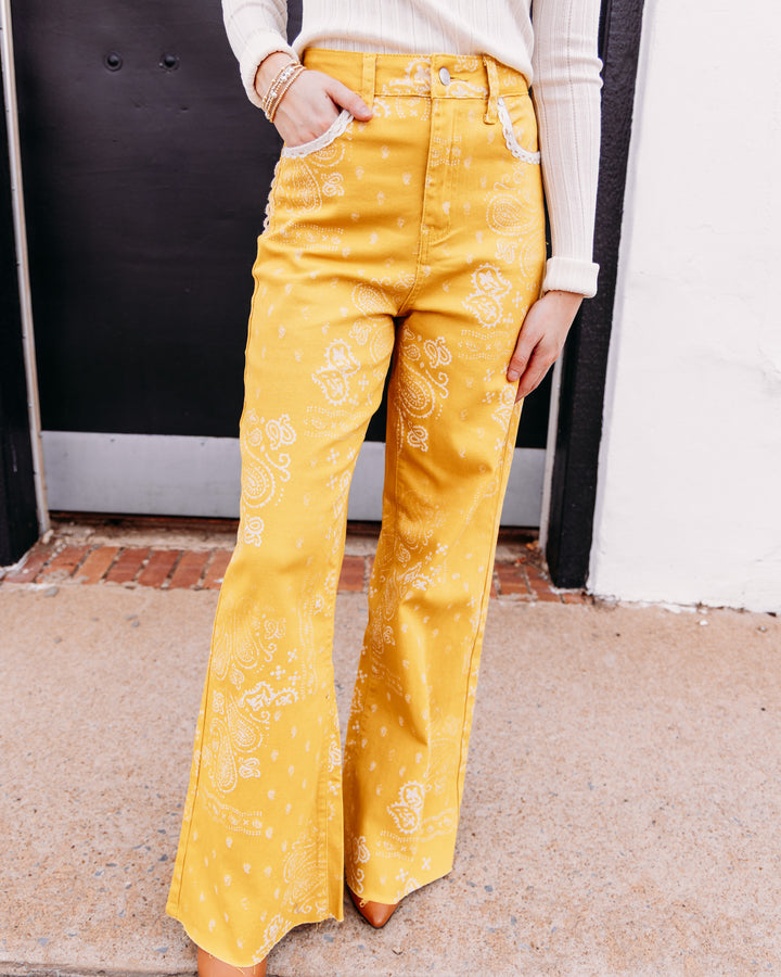 A person wearing bright yellow wide-leg pants with a white paisley bandana print, paired with a cream ribbed long-sleeve top. The photo is cropped from the chest down, showing the pants in a lifestyle setting against a black and white door.