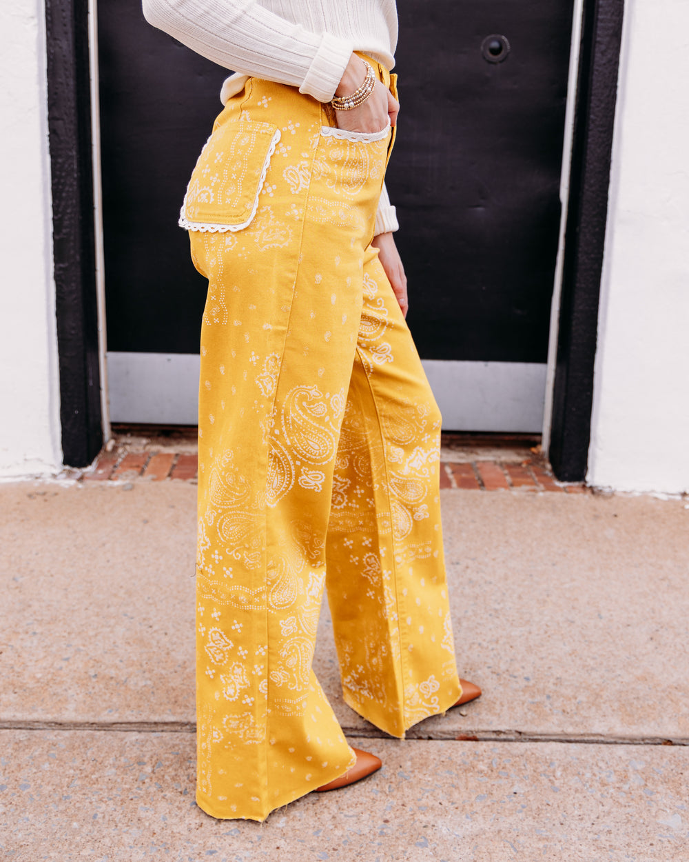 A person wearing bright yellow wide-leg pants with a white paisley bandana print, lace trim along the pocket edges, and a white ribbed top, standing on a sidewalk in front of a black door.