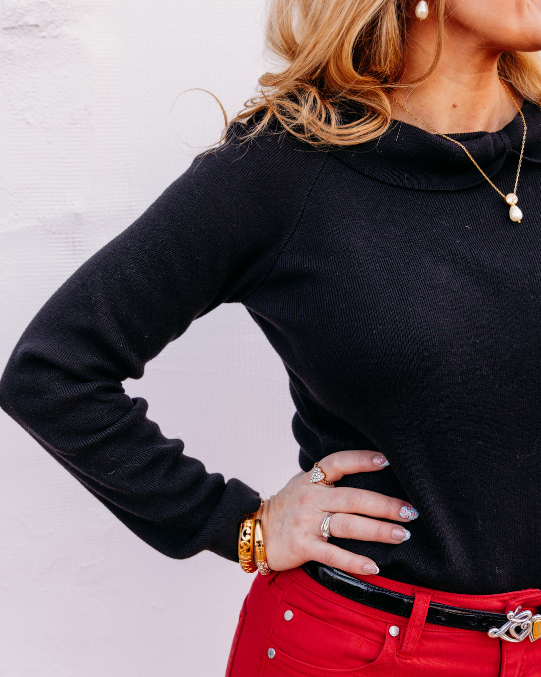 A woman wearing a black ribbed long-sleeve mock neck top paired with bright red pants and gold jewelry, posing with one hand on her hip against a light textured wall.
