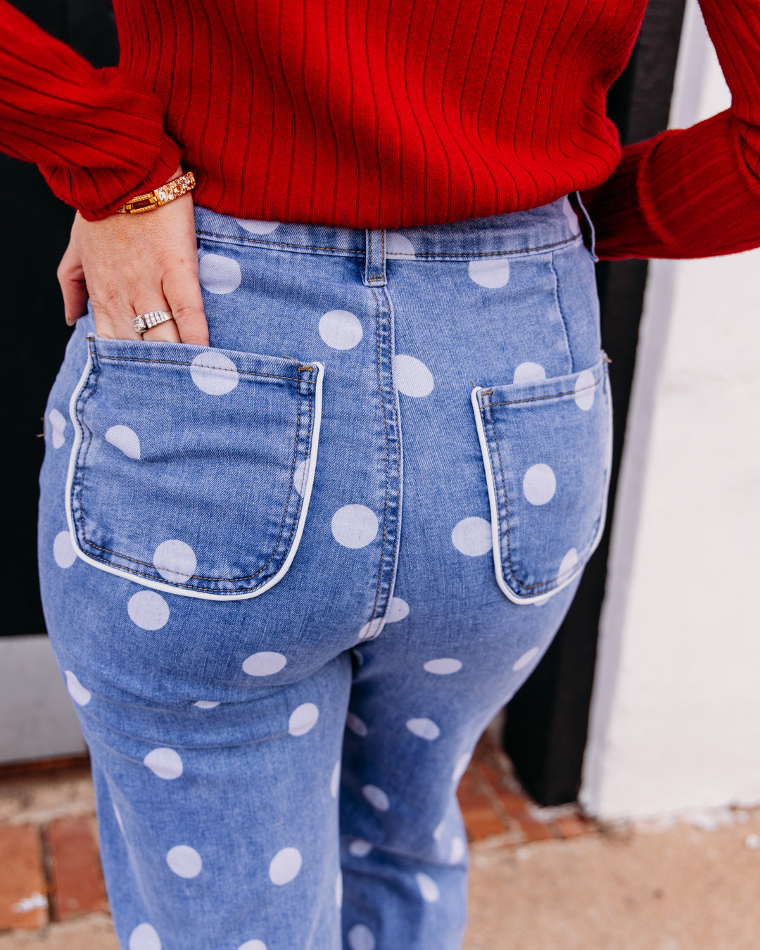 A close-up back view of a person wearing blue denim jeans with large white polka dots and a red ribbed sweater, standing outdoors near a doorway.