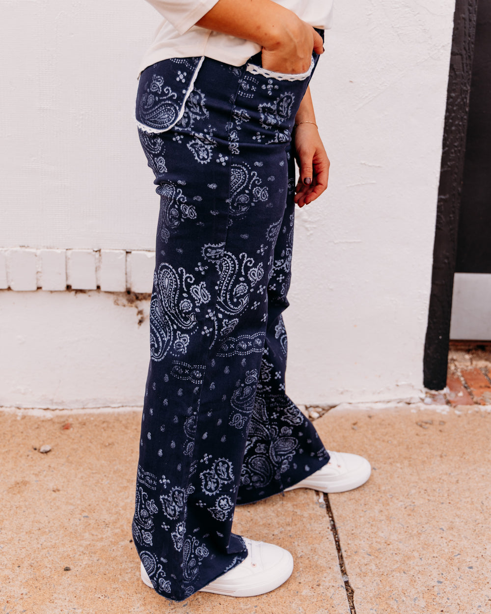 A person wearing navy blue wide-leg pants with a white paisley bandana pattern, styled with a cream top and white sneakers, standing against a white stucco wall.