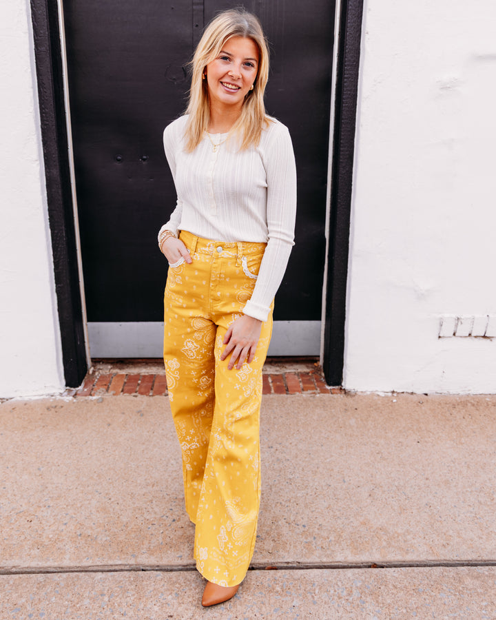 A woman stands outdoors in front of a white wall and black door, wearing a cream ribbed long-sleeve top and bright yellow wide-leg pants with a white floral pattern.
