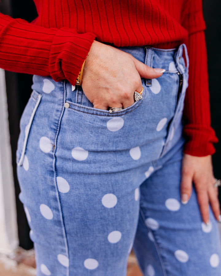 Close-up of a person wearing light blue denim jeans with large white polka dots, paired with a red ribbed sweater. The person's hand is in the jeans' front pocket.