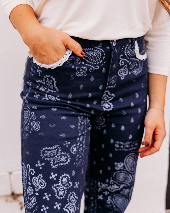 A close-up of a person wearing navy blue jeans with a white paisley bandana print and white lace trim along the pocket edges, paired with a white long-sleeve top.