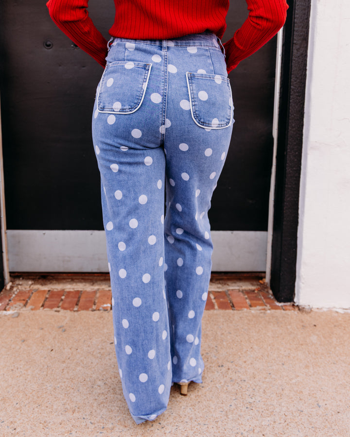 A person viewed from behind wearing light blue wide-leg jeans with large white polka dots, paired with a red ribbed sweater, standing outdoors in front of a dark door.