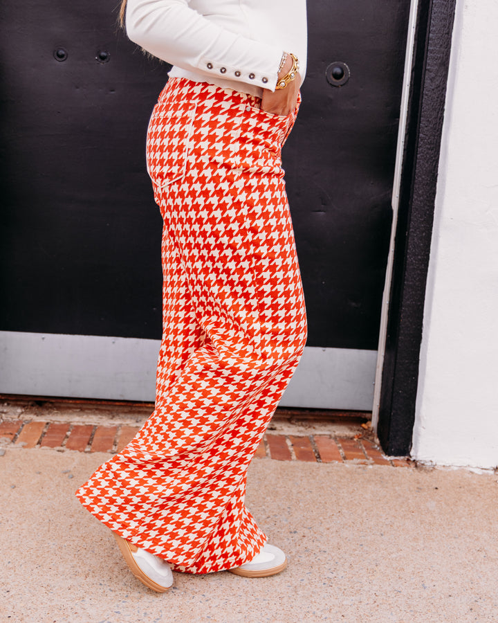 A person wearing bright orange and white houndstooth wide-leg pants, paired with a white long-sleeve top and white shoes, standing in front of a black door.
