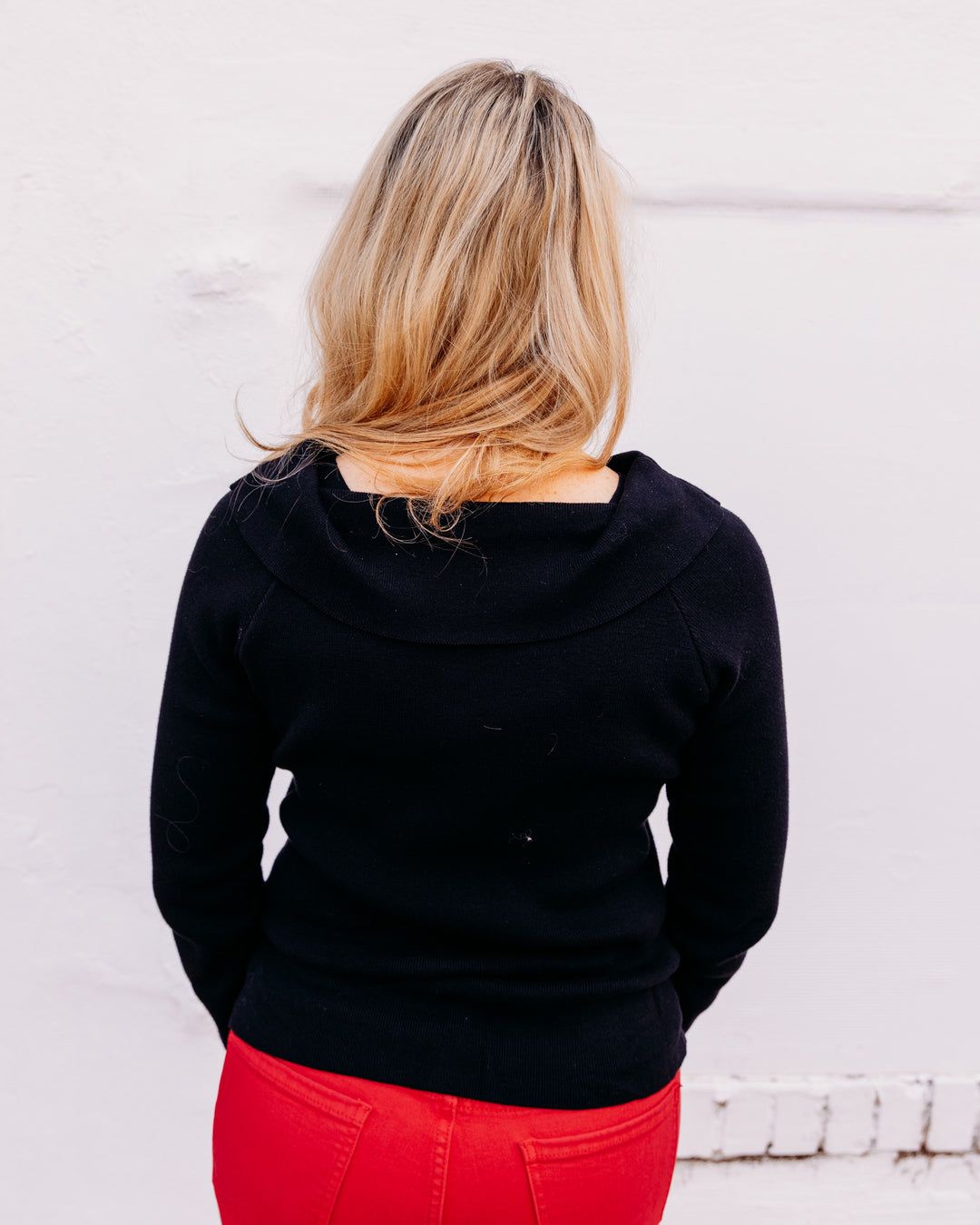 A woman viewed from behind wearing a black long-sleeve top with a wide draped neckline and bright red pants, standing against a white wall.