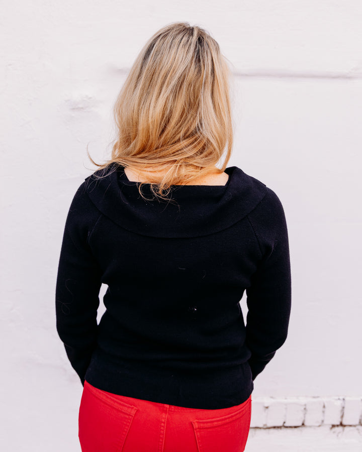 A woman viewed from behind wearing a black long-sleeve top with a wide draped neckline and bright red pants, standing against a white wall.