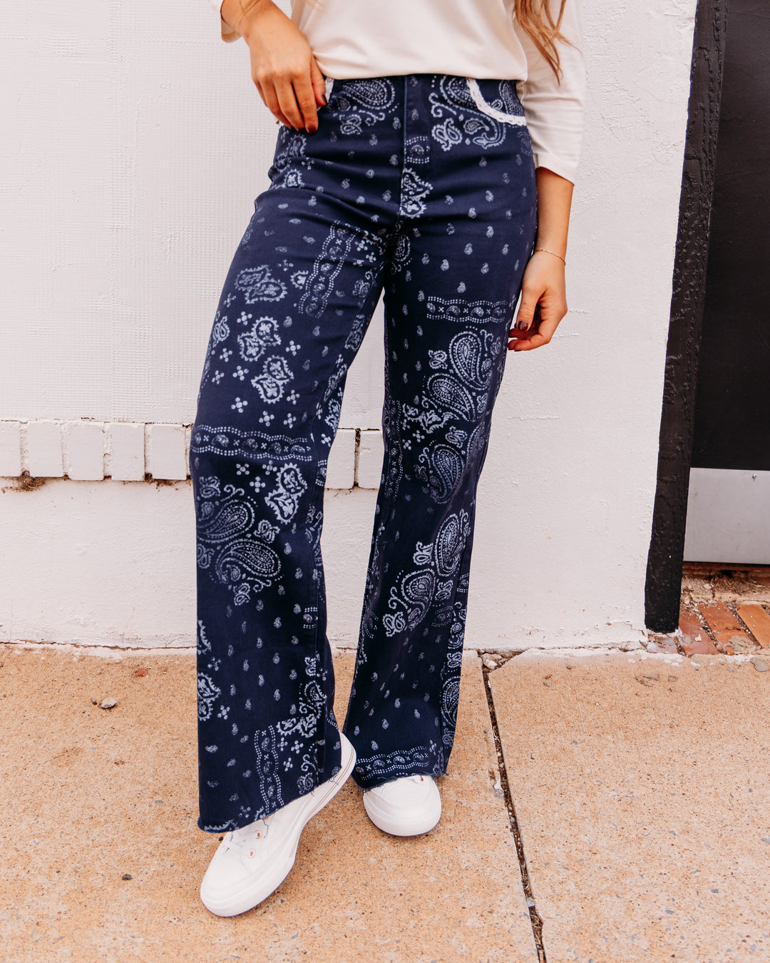 A person wearing dark navy wide-leg jeans with a white paisley bandana print, styled with a cream long-sleeve top and white sneakers, standing against a white wall.