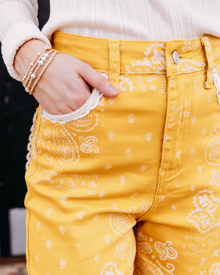 Close-up of a person wearing mustard yellow jeans with a white paisley bandana print, a cream ribbed sweater, and layered gold bracelets.