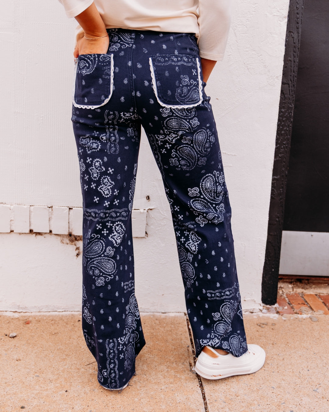 A person shown from the back wearing navy blue wide-leg pants with a white paisley bandana pattern, a cream top, and white sneakers, standing outdoors against a white wall.