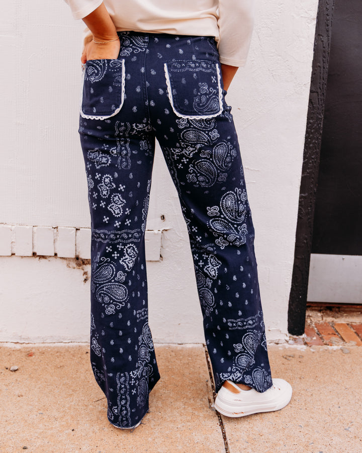 A person shown from the back wearing navy blue wide-leg pants with a white paisley bandana pattern, a cream top, and white sneakers, standing outdoors against a white wall.