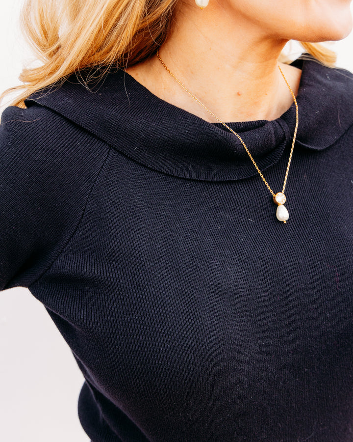 A close-up lifestyle shot of a woman wearing a delicate gold chain necklace with a double pearl pendant, paired with a dark navy ribbed knit top.